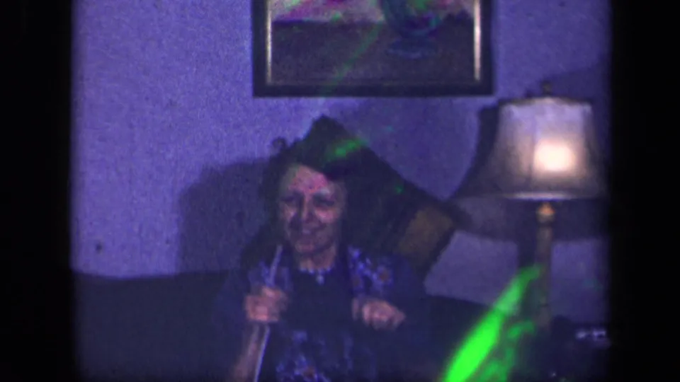 The woman smiles toward the camera while holding knitting needles in a dim Sebring, Ohio living room.