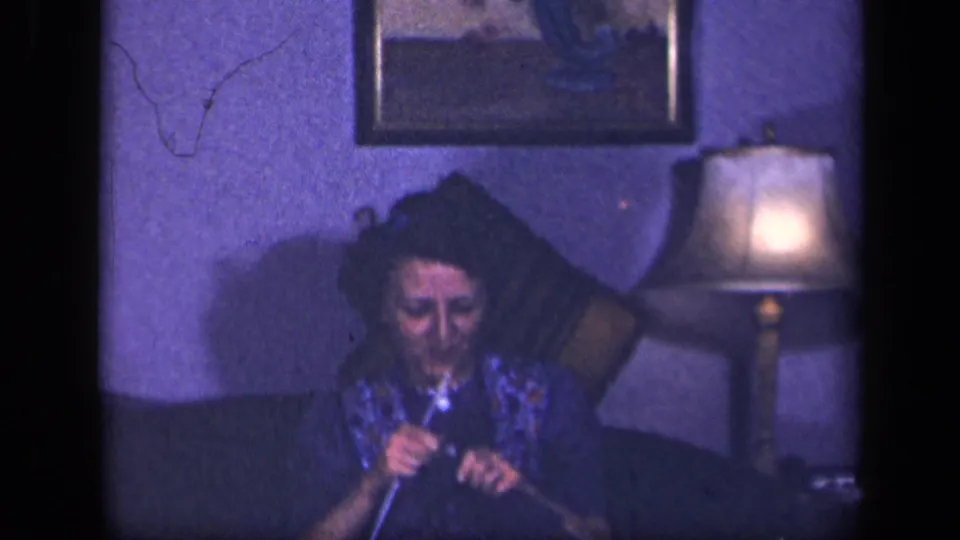 A woman in a floral dress looks down at her knitting in a softly lit 1940s home interior.