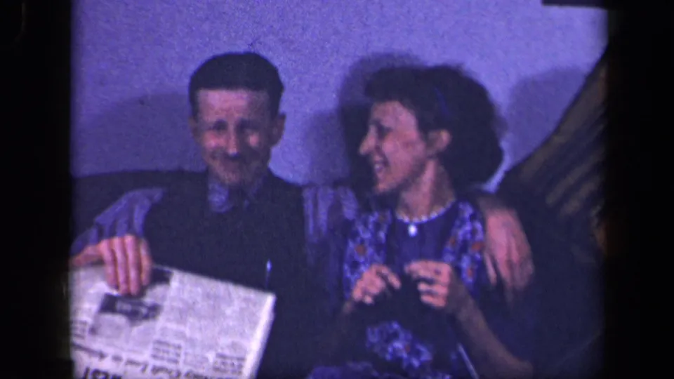 A man sits beside the crocheting woman in the 1942 Sebring living room, smiling as he holds a newspaper on the couch.