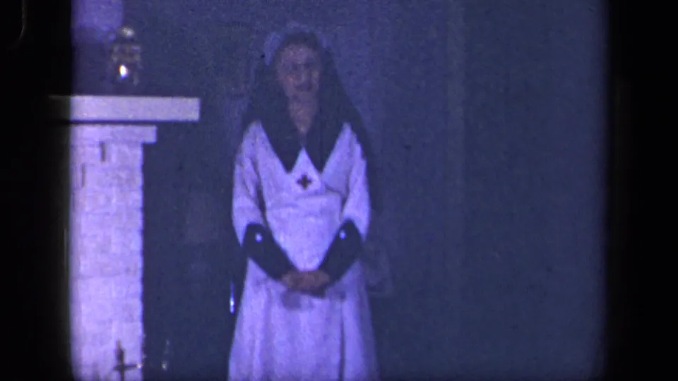A soft 8mm frame shows the nurse-costumed woman standing near the fireplace, with blue-purple tint and low interior light.