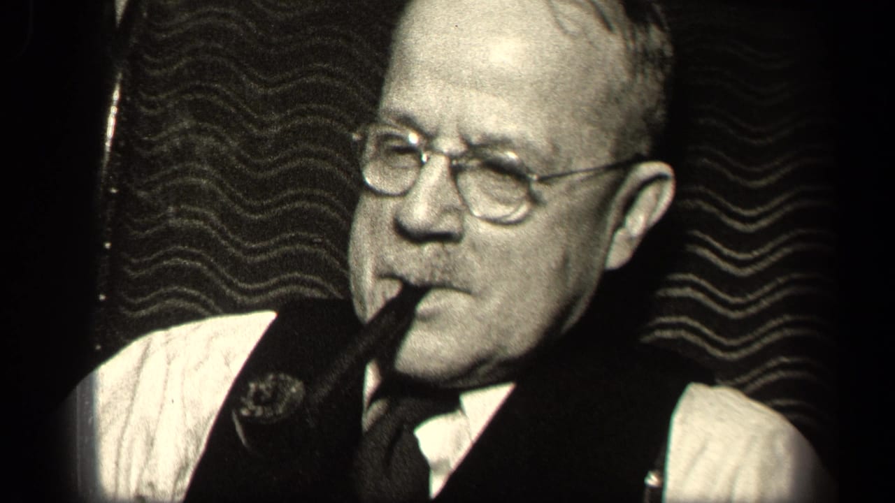 Older man in round glasses holds a pipe in his mouth against a dark patterned background.