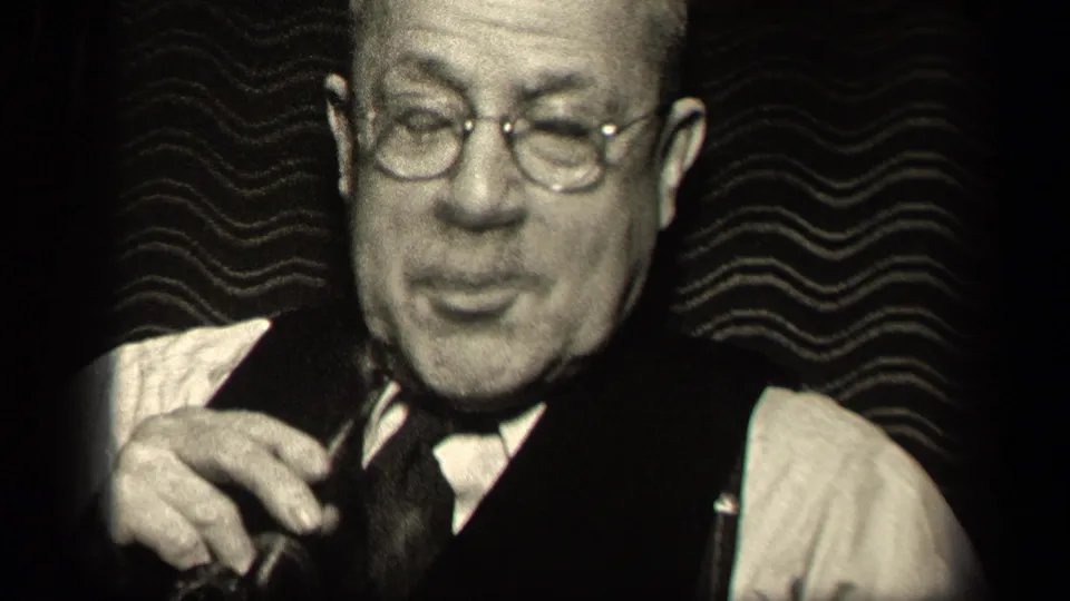 1942 Sebring, Ohio frame showing an older man with glasses lowering a pipe while seated against a patterned background.