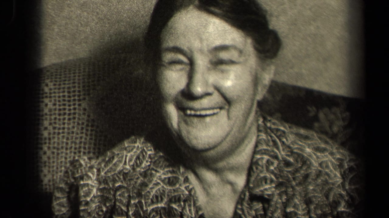 Older woman in a patterned blouse smiles while seated indoors.