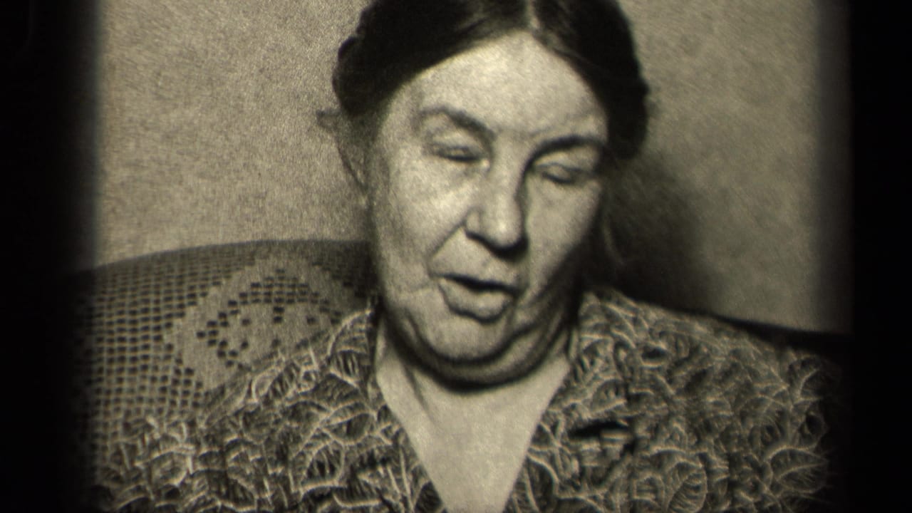 Older woman seated indoors with her mouth partly open as if speaking.