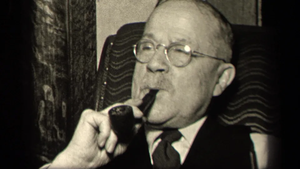 The older man remains seated in the patterned chair, wearing round glasses and a suit as he draws on a pipe in the 1942 Sebring, Ohio interior footage.