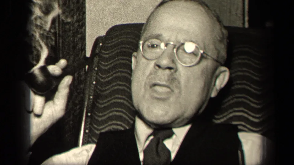 A close home-movie view shows the suited older man with round glasses holding his pipe away from his mouth as smoke rises in the dim interior.