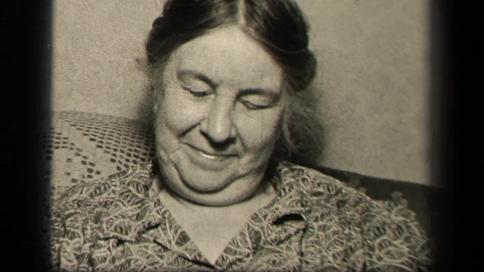 An older woman in a patterned blouse sits indoors on a chair with a crocheted covering, smiling gently downward in the Sebring, Ohio home-movie footage.