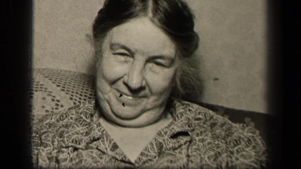 In the 1942 Sebring, Ohio home-movie frame, an elderly woman in a patterned blouse smiles toward the camera while seated against an upholstered back and plain wall.