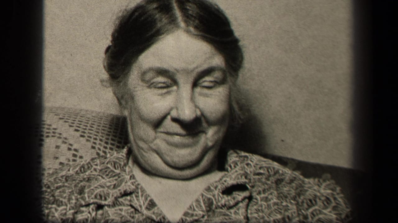 Older woman seated indoors, smiling toward the camera beside part of another seated woman.