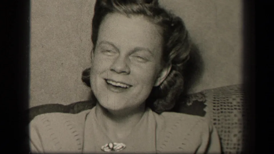 In this 1942 Sebring, Ohio home-movie frame, a young woman in a light dress and brooch smiles while seated on patterned upholstery.