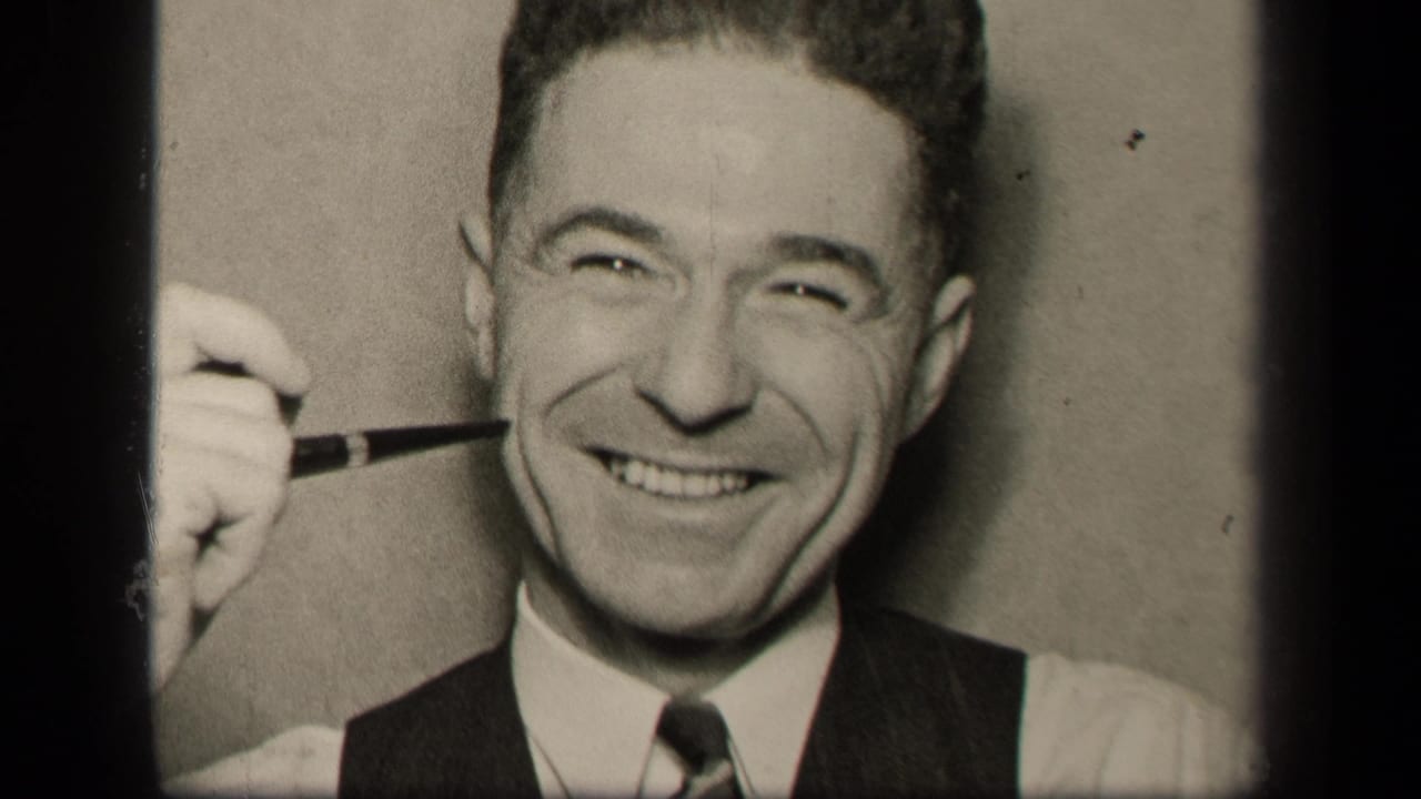Smiling adult man in a shirt, tie, and vest holds a pipe beside his face.