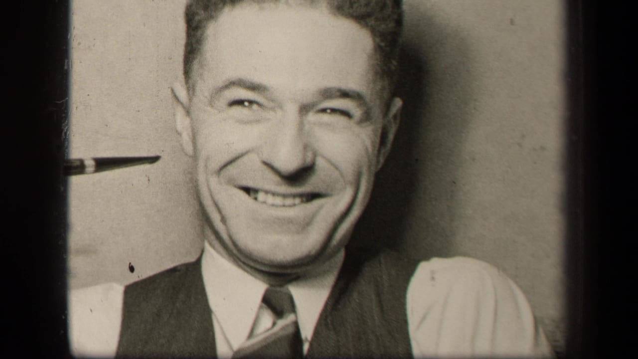 Man in vest and tie smiles broadly while seated indoors near a pipe.