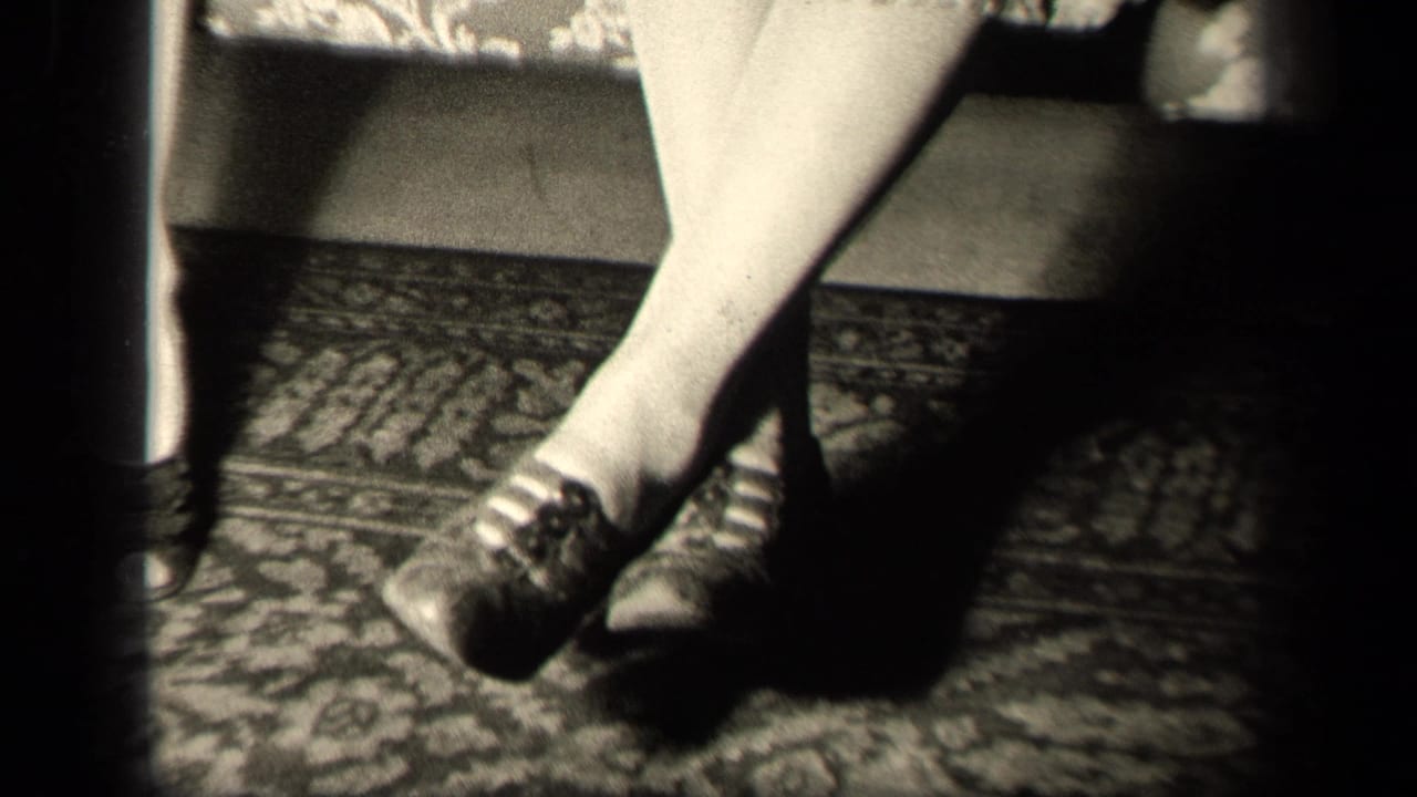 1942 Sebring, Ohio home-movie frame showing a seated woman's crossed legs, stockings, dark lace-up shoes, floral couch, and rug.