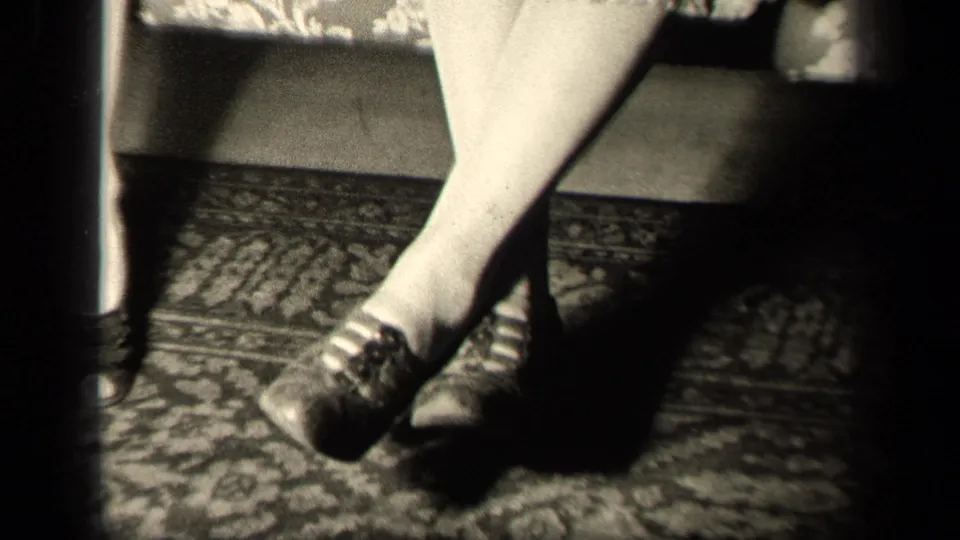 1942 Sebring, Ohio home-movie frame showing a seated woman's crossed legs, stockings, dark lace-up shoes, floral couch, and rug.