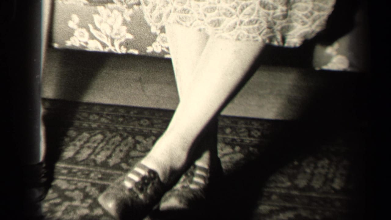 Close 1942 Sebring, Ohio frame of a seated woman's legs with stockings, dark shoes, a lace dress hem, and floral couch upholstery.