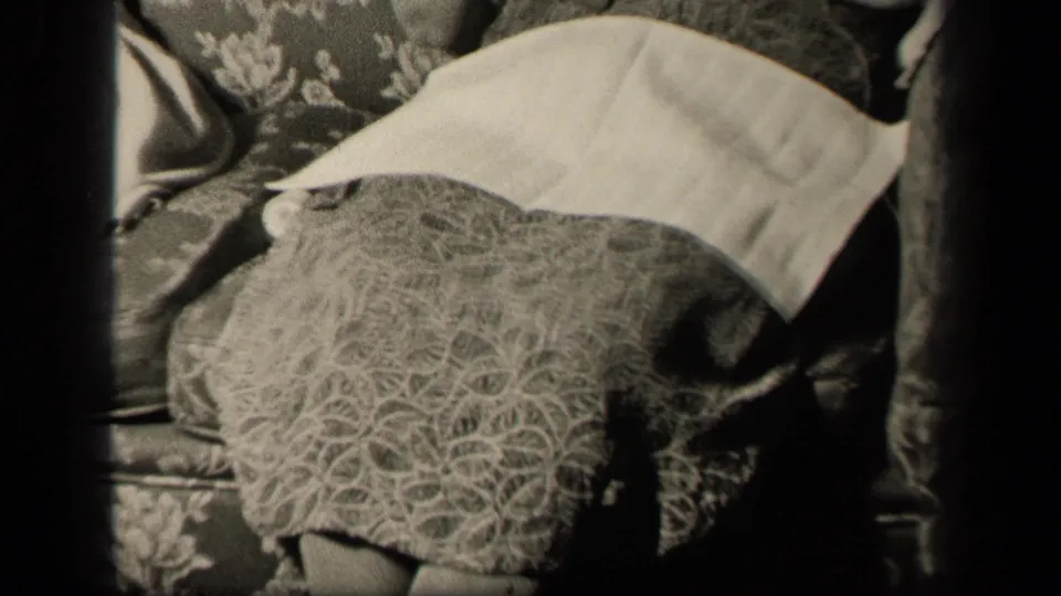 Angled 1942 Sebring, Ohio home-movie frame of a seated woman’s patterned skirt, crossed legs, lap napkin, and floral couch.