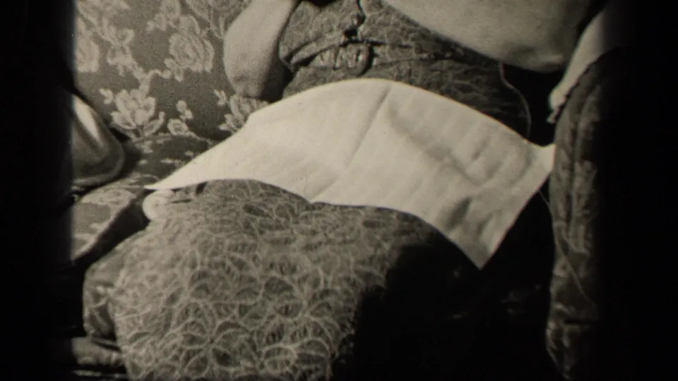 1942 Sebring, Ohio frame with the seated woman partly cropped, showing her lap napkin, patterned skirt, couch upholstery, and nearby furniture.