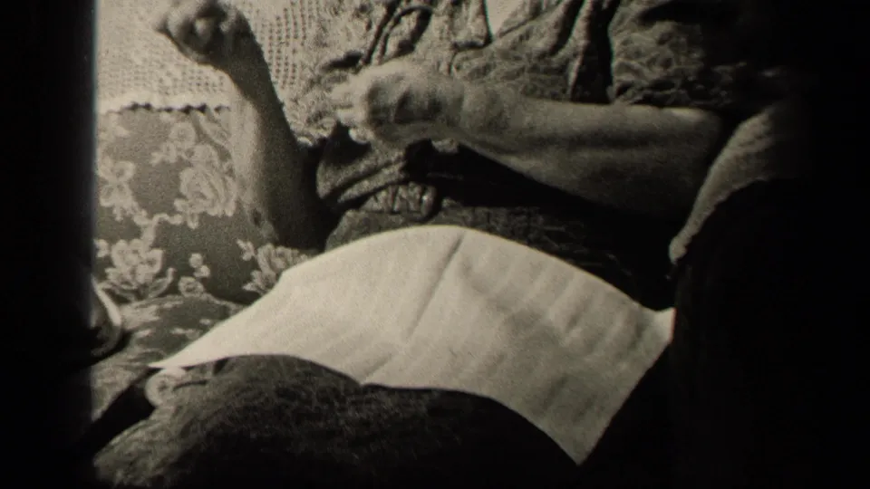A seated woman in a dark dress and white apron pauses or adjusts her knitting on a floral couch in the supplied 1942 Ohio home-movie setting.