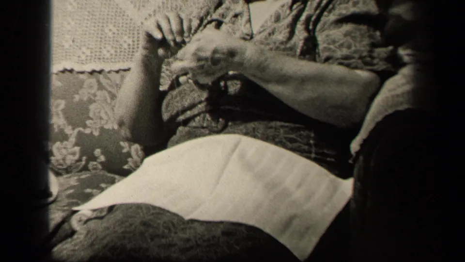 The final frame centers on the woman's hands and lap as she knits, with the floral couch and soft black-and-white home-movie texture visible.