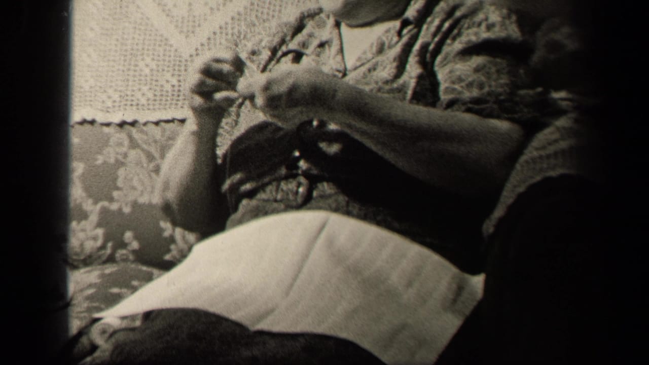 Older woman in a 1942 Sebring, Ohio home interior sits on a patterned couch while handling needlework near her lap.