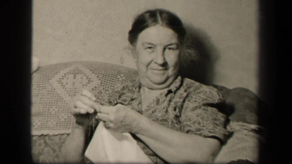 Older woman in the 1942 Sebring, Ohio home movie smiles toward the camera while holding needlework on the couch.