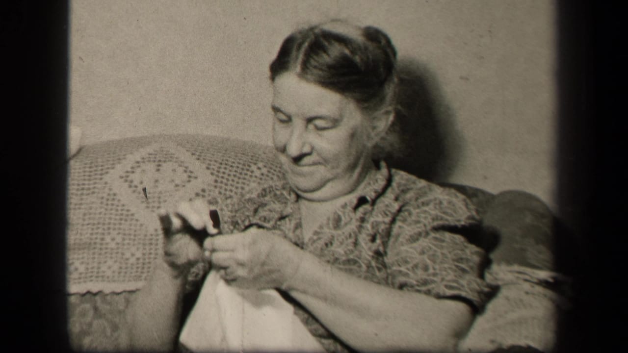 Older woman seated on a couch sewing a white cloth while looking down.
