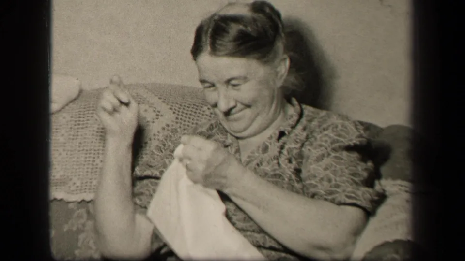 An older woman smiles down at a white cloth as she continues sewing on a couch in metadata-supported 1942 Sebring, Ohio.