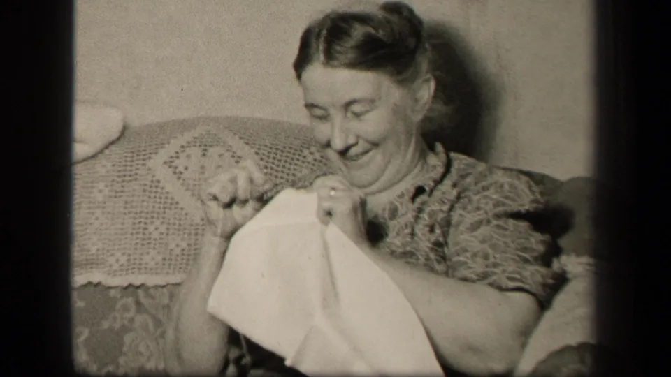 The seated woman lifts the white cloth while sewing, framed closely in black-and-white 8mm footage from Sebring, Ohio in 1942.