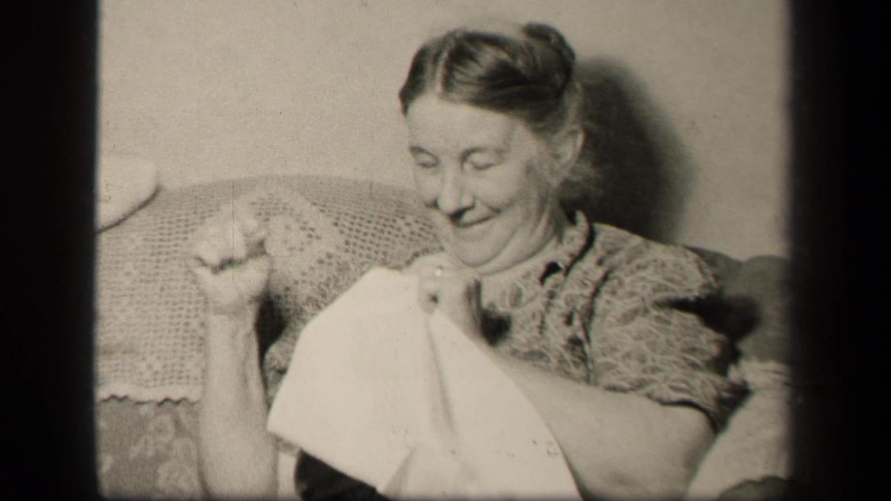 Woman seated in a floral armchair sewing fabric and smiling downward.