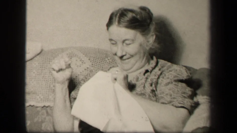 In 1942 Sebring, Ohio home-movie footage, a woman sits in a floral armchair sewing or mending fabric.