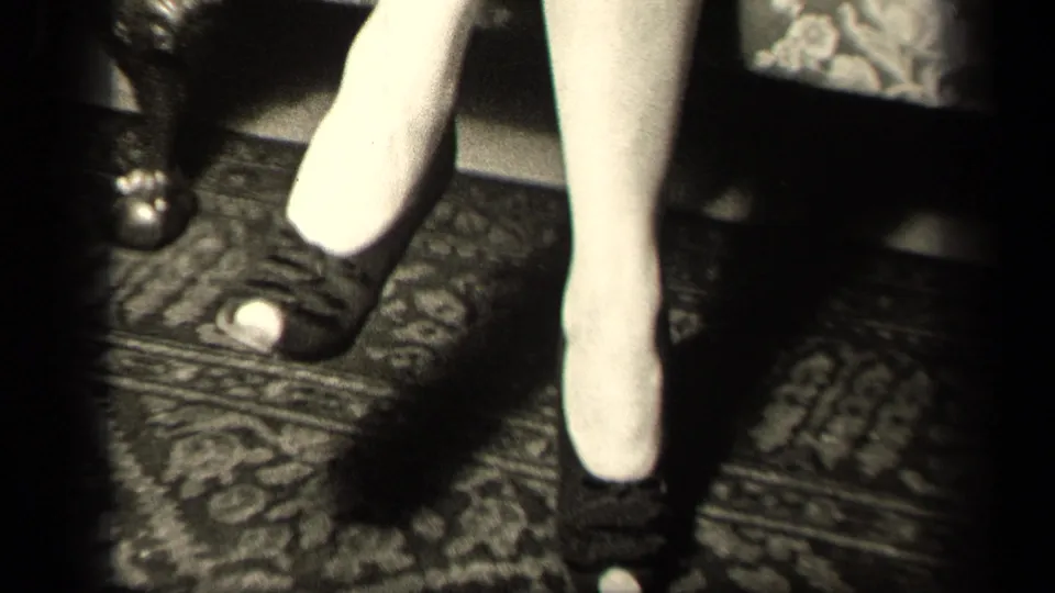 Low-angle 8mm frame from the Sebring, Ohio interior focuses on the woman's stockings and dark heels near the armchair.