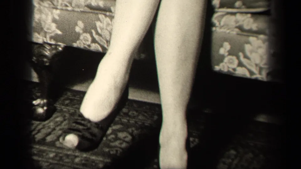 Close home-movie view shows the seated woman's legs, white stockings, and one dark high heel beside floral upholstery.