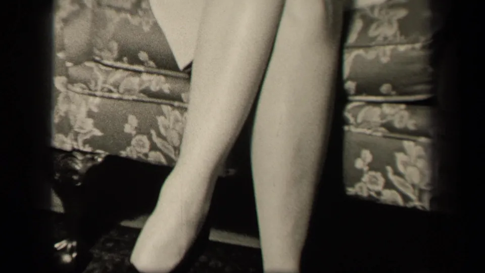 The final frame holds on the seated woman's white-stockinged legs against the floral armchair in the private interior.