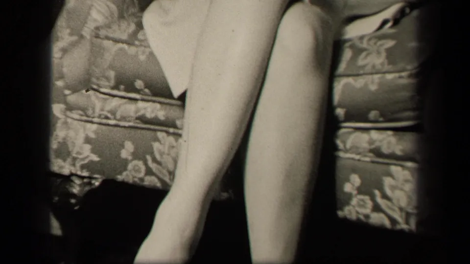 1942 Sebring, Ohio home-movie frame showing a seated woman's crossed legs, stockings, light dress, and floral couch upholstery.