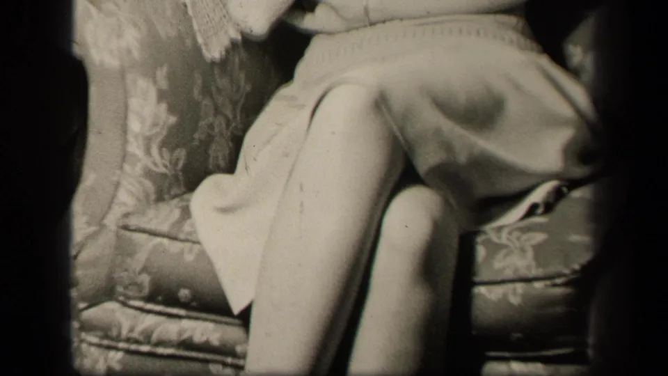 1942 Sebring, Ohio home-movie frame showing the seated woman's lap, folded legs, light dress, and floral upholstered couch.