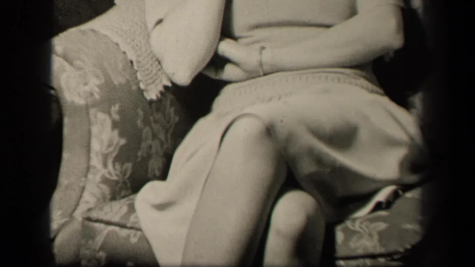 1942 Sebring, Ohio frame showing the seated woman's torso, lap, bracelet, light dress, crossed legs, and couch upholstery.