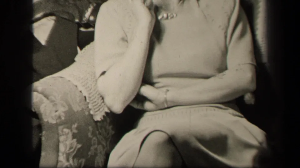 The camera continues upward on the seated woman in the ornate armchair, with her cardigan sleeve and lap still prominent.