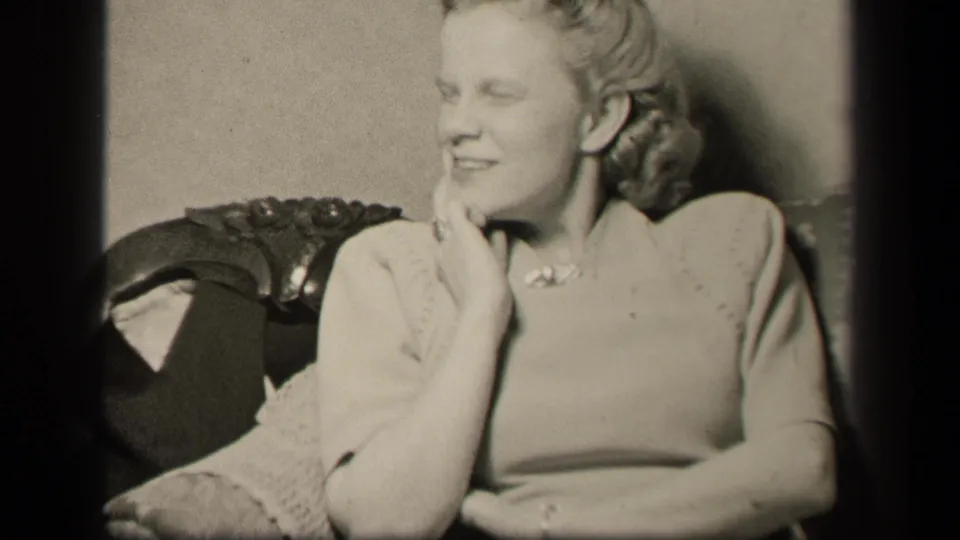 In 1942 home-movie footage, the seated woman turns slightly and holds her hand near her chin in an informal interior portrait.