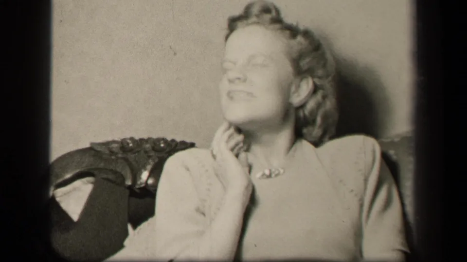 In 1942 Sebring, Ohio home-movie footage, a woman in a light dress sits in an ornate chair, eyes partly closed, hand raised near her neck.