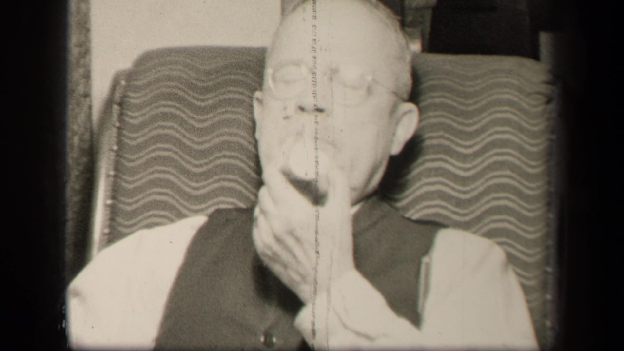 Older man in glasses and vest sits in a patterned chair while holding fruit to his mouth.