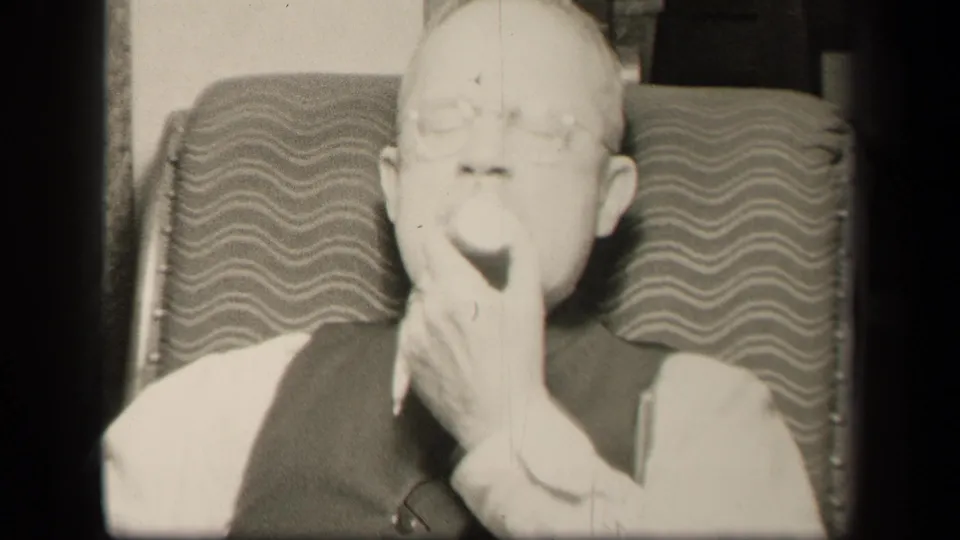 1942 home-movie frame of the older man seated indoors, holding fruit near his mouth against the patterned chair back.