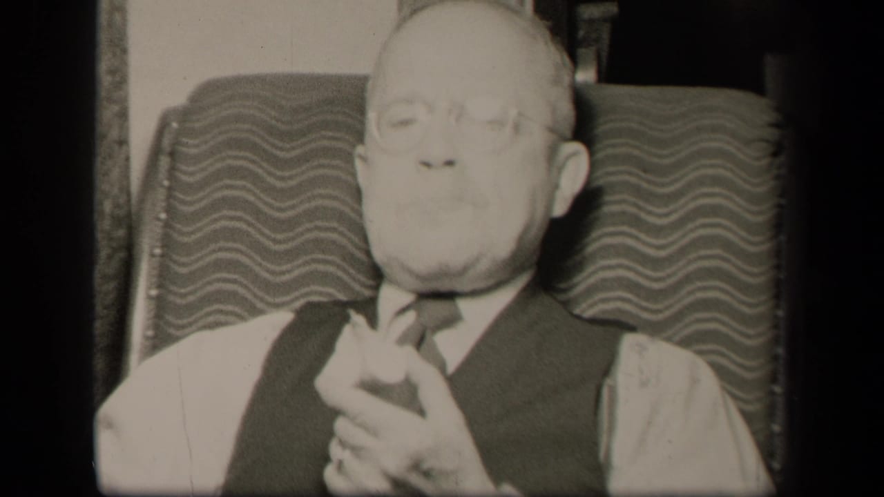 Elderly man with glasses seated in a patterned easy chair, hands clasped near his chest.
