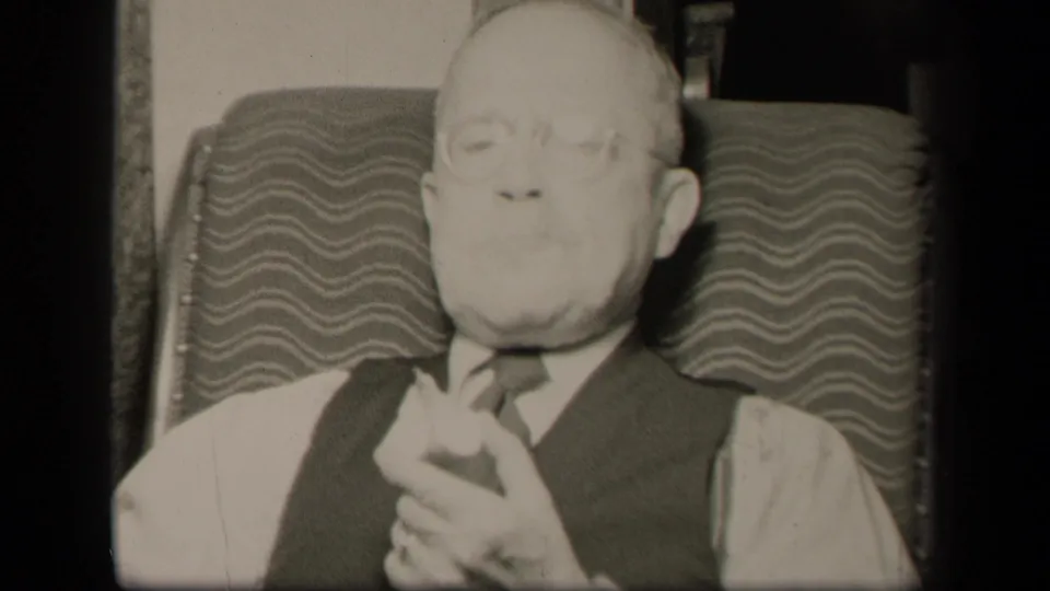 In a 1942 Sebring, Ohio home-movie frame, an elderly man in glasses, vest and tie sits in a patterned easy chair with hands clasped.