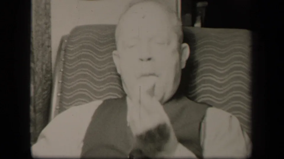 A close 1942 home-movie frame shows the elderly man seated in the patterned chair with his hand near his chin and eyes lowered.