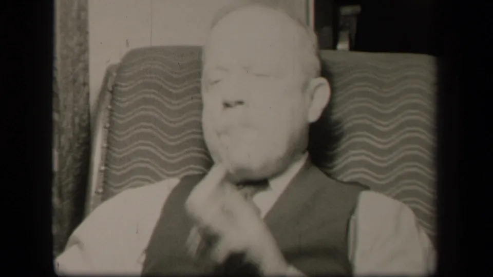 The final frame shows the elderly man still seated in the patterned easy chair, hand near his mouth, in monochrome 8mm footage.