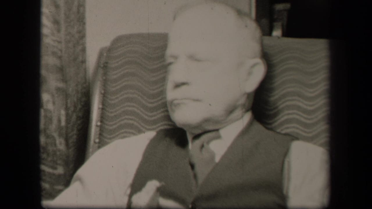 Older man in vest and tie seated in a patterned armchair, turned slightly to the side.