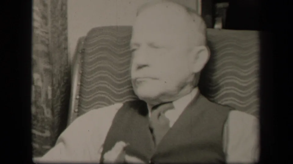 In this 1942 Sebring, Ohio home-movie frame, an older man sits in a patterned armchair wearing a light shirt, dark vest, and tie.