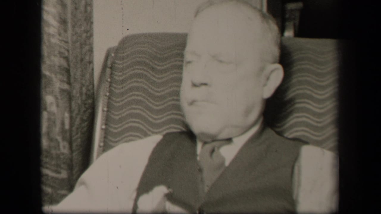 The seated man appears in soft-focus 1942 home-movie footage, facing forward in a patterned chair inside a private room.