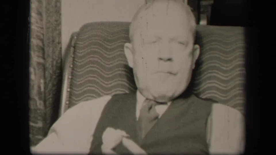 A closer forward-facing view shows the older man seated in the patterned armchair, with his hand partly visible near his clothing.