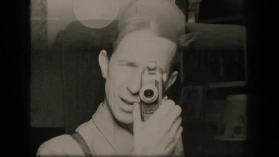 A younger man, likely the filmmaker, films his mirror reflection with a small 8mm camera inside the same private interior.